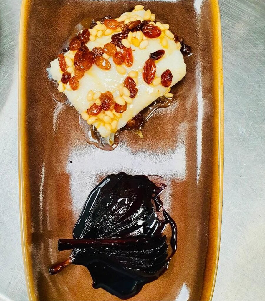 Bacallà amb salsa agredolça de mel, panses i pinyons sobre una base de ceba caramel·litzada